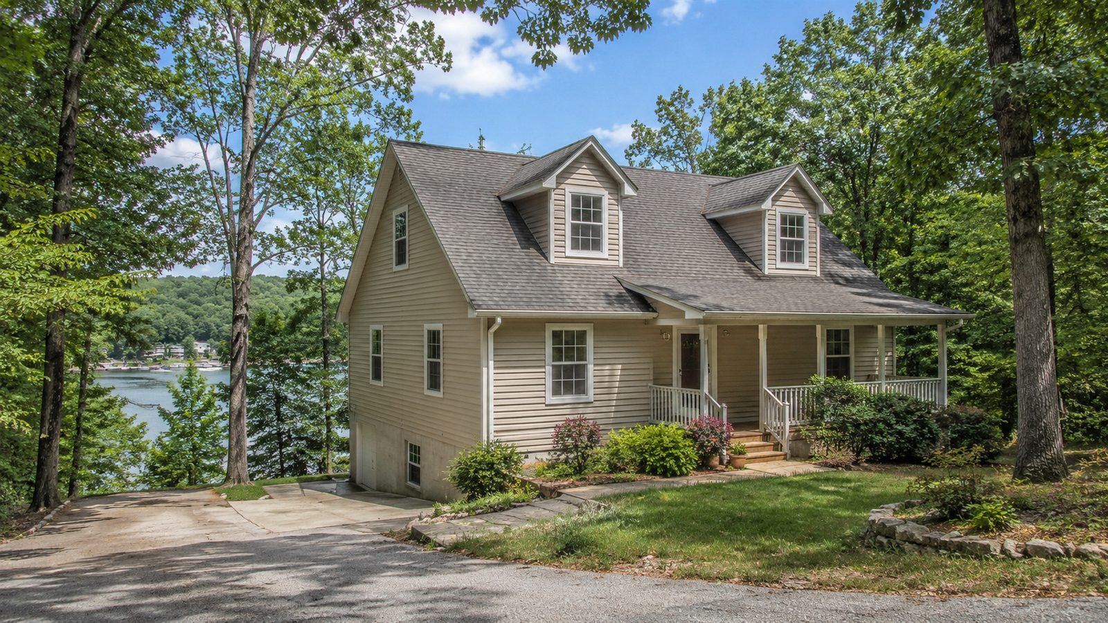 A two-storey clapboard house with dormer windows and a covered front porch, set among mature oaks on a sloping wooded lot, with a cove of the lake and a dock visible through the trees behind it.